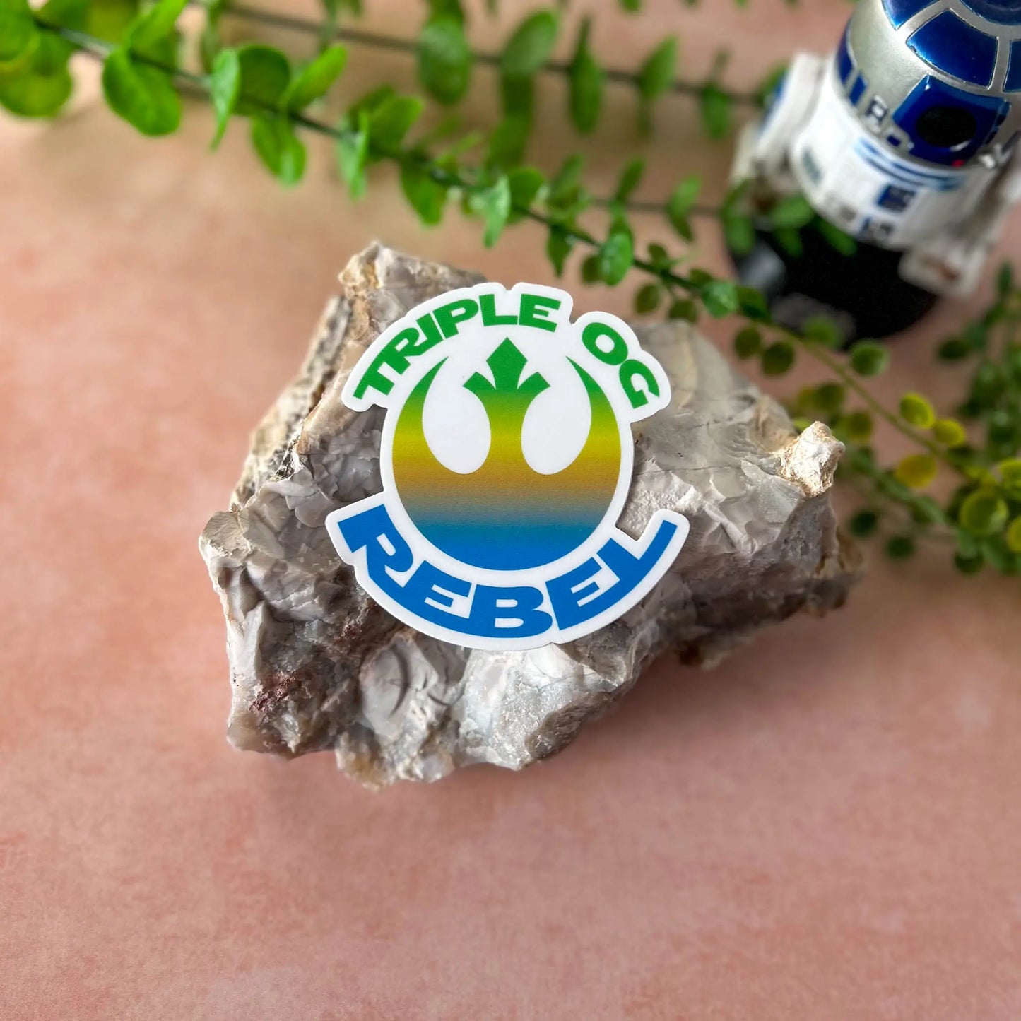 Triple OG Rebel 3" Vinyl Sticker on a rock with foliage and an R2-D2 bobblehead in the background