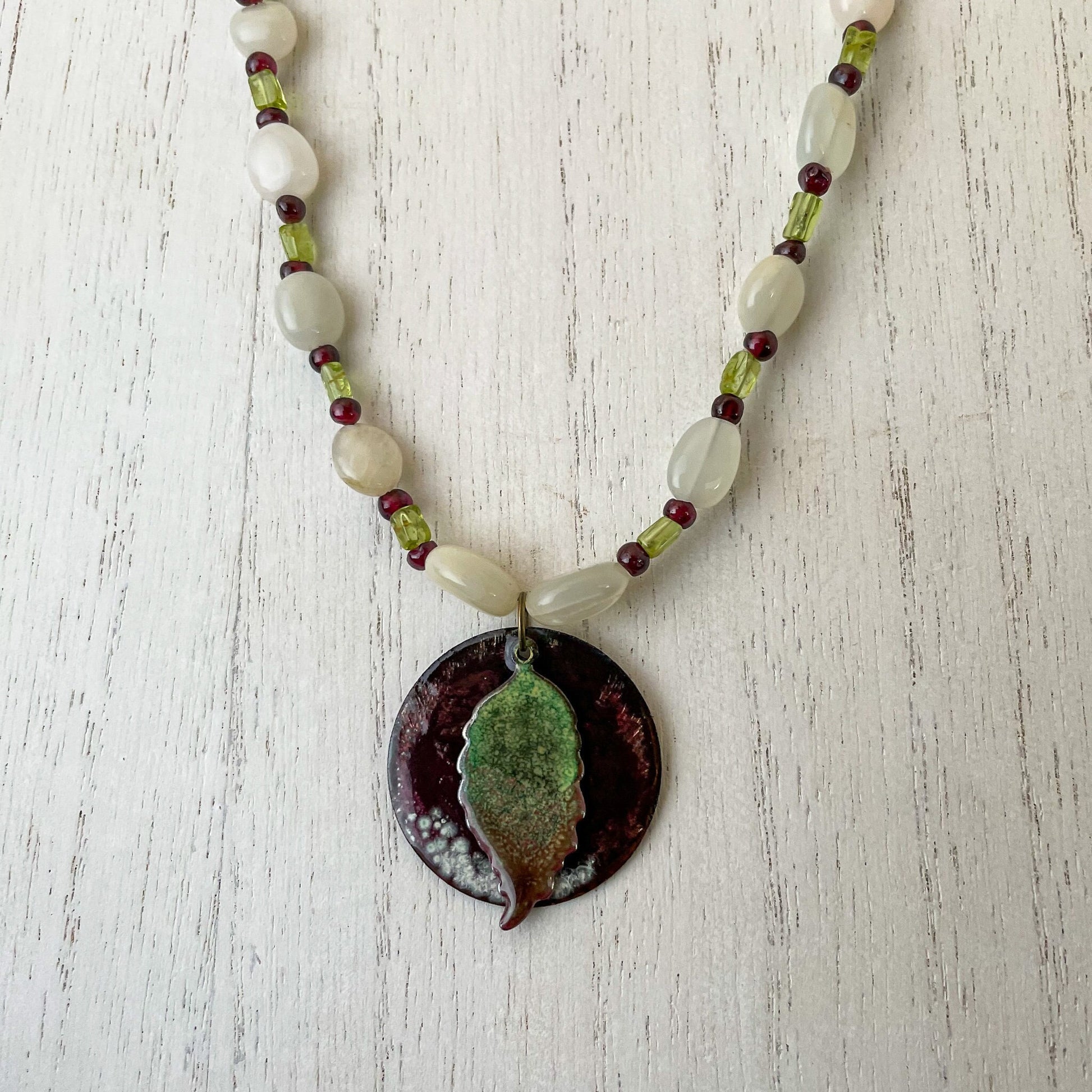 Green Leaf and Burgundy Circle Layered Enamel Pendant Moonstone, Garnet, and Peridot Bead Necklace on white wood
